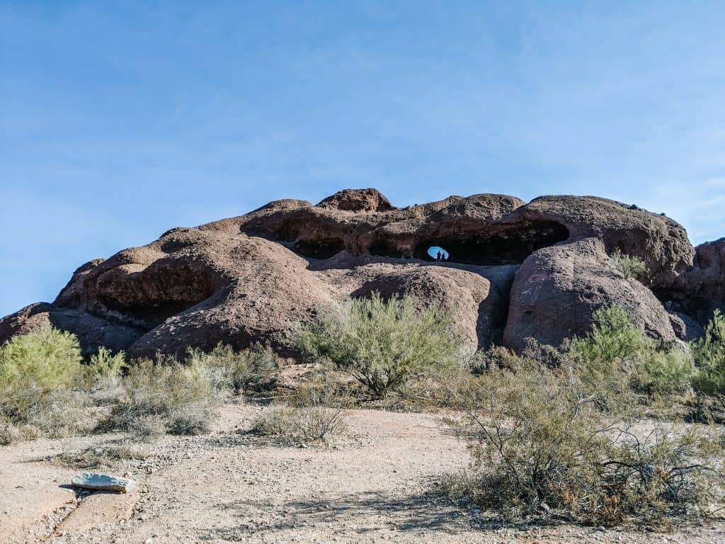 A unique rock formation with natural openings, seen during a hike in Phoenix. The rugged, reddish-brown rocks are contrasted against the clear blue sky, with thin desert vegetation in the foreground. This spot is a popular destination for those hiking in Phoenix, offering stunning views and interesting geological features.