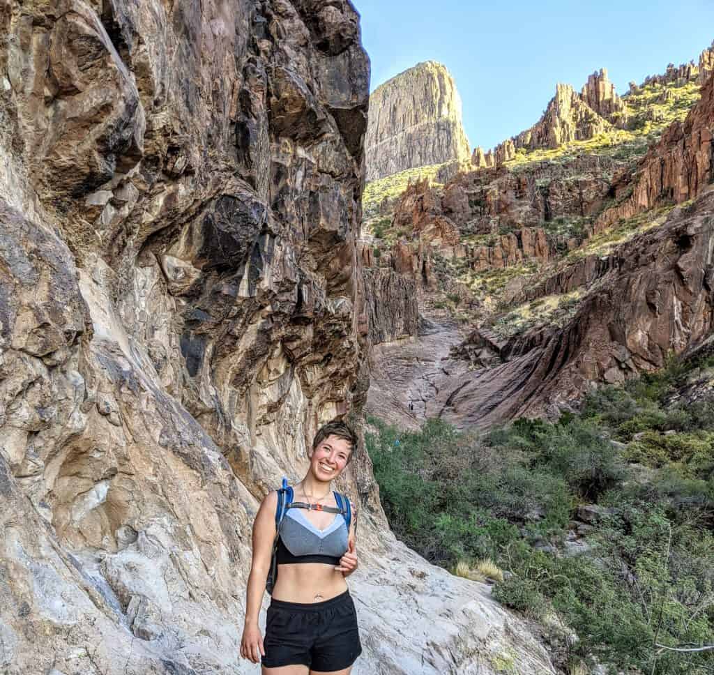 I paused for a photo against a rocky cliff with the bright sunlight illuminating the rugged terrain behind me. The steep canyon walls and lush greenery in the background highlight the diverse and beautiful scenery you can experience while hiking in Phoenix.