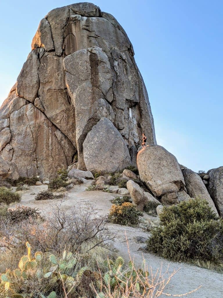 A large rock formation stands tall against the clear blue sky with a hiker standing near its base, highlighting the dramatic and rugged terrain typical of hiking in Phoenix. Cacti and desert shrubs populate the sandy trail leading up to the imposing structure, capturing the essence of the Sonoran Desert landscape.