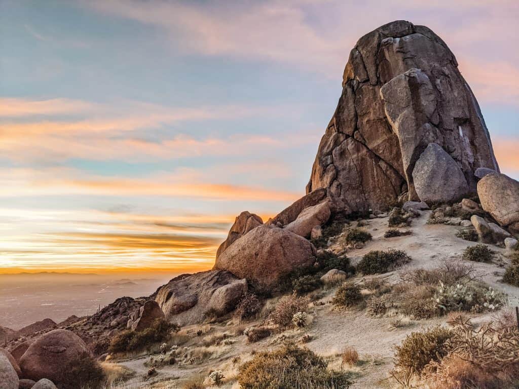 A stunning sunset behind a towering rock formation, surrounded by desert vegetation, highlighting the serene beauty of hiking in Phoenix.