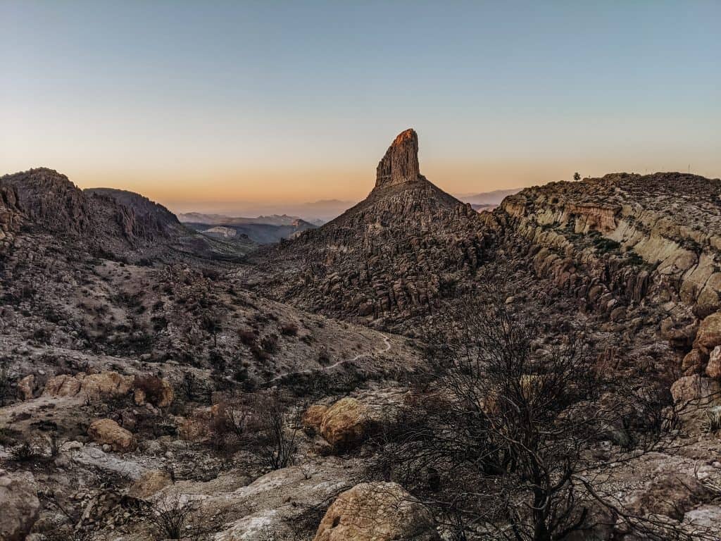 A rock formation rising prominently from a rocky desert landscape, captured during sunset. The sky is softly lit with pastel hues, highlighting the serene and rugged beauty typical of hiking in Phoenix.