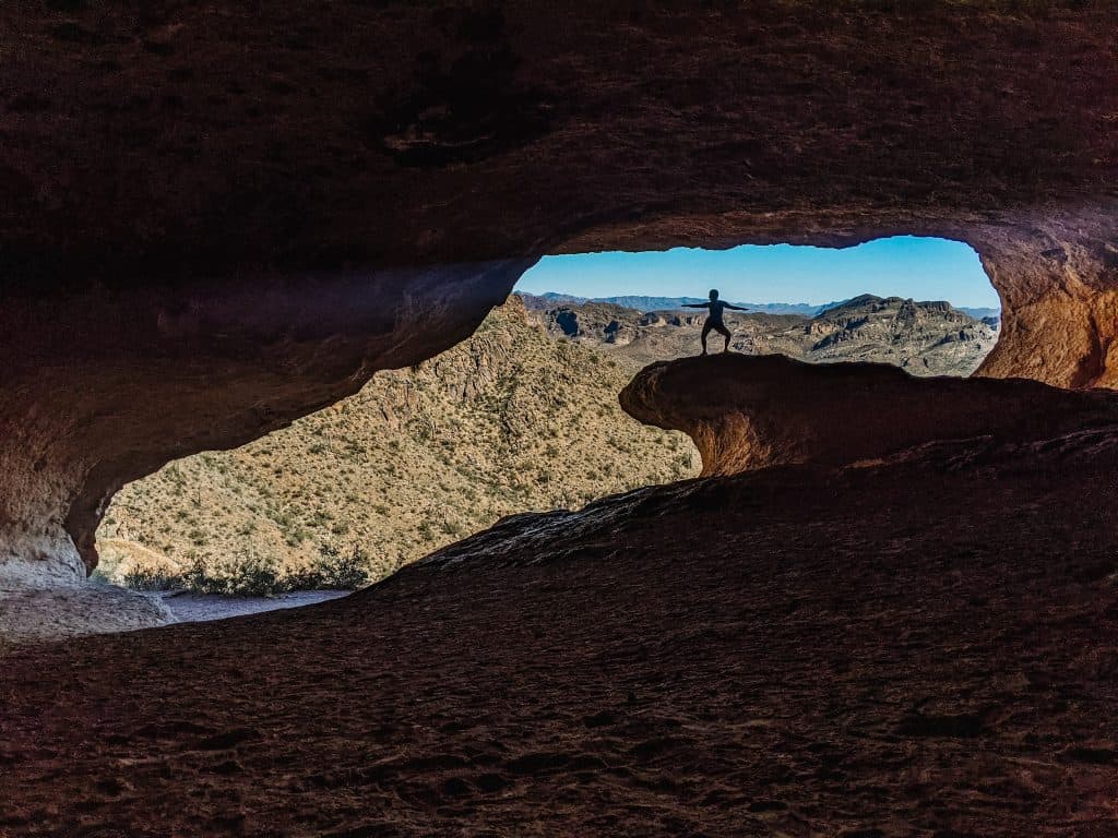 Standing on a rocky outcrop inside a large cave, overlooking the desert landscape and mountains while hiking in Phoenix.