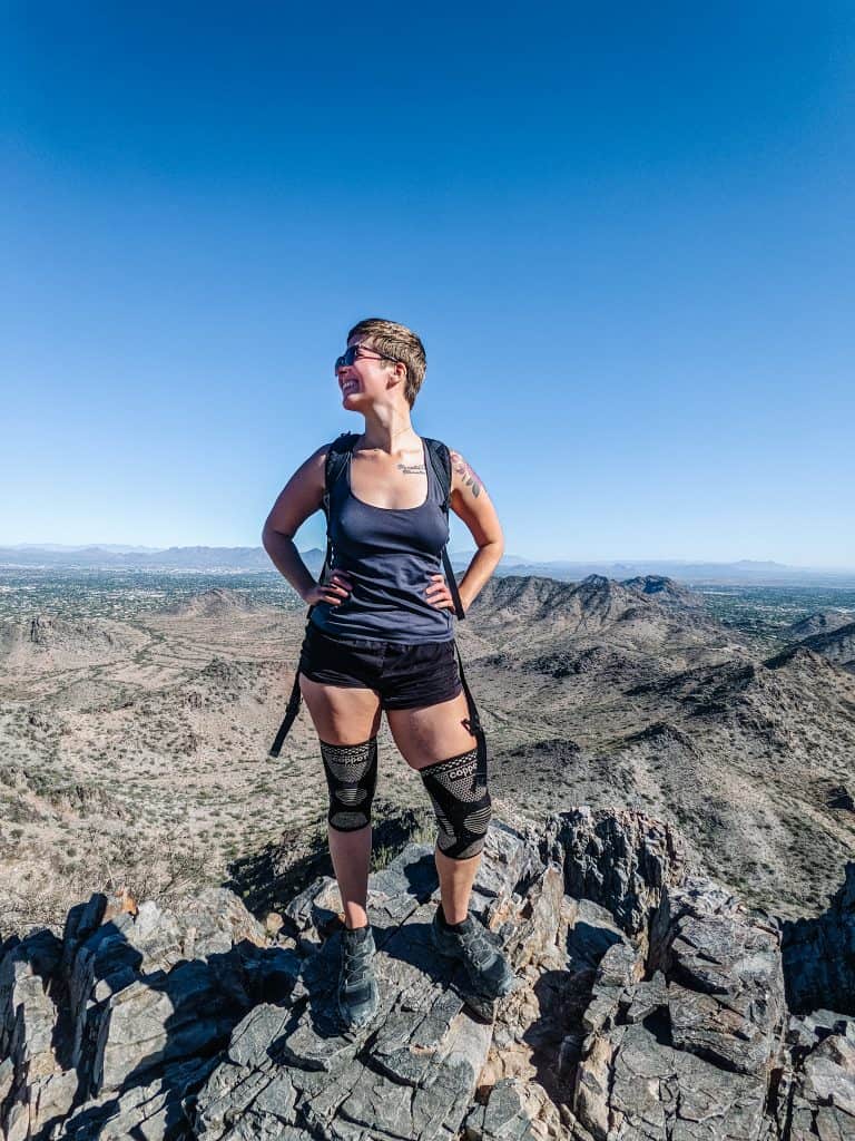 I'm standing confidently on a rocky peak, with the expansive desert landscape of Phoenix stretching out behind me under a clear blue sky while hiking in Phoenix.