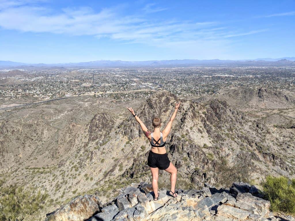 I’m standing on a rocky peak with my arms raised in celebration, overlooking the desert landscape and cityscape of Phoenix. The clear blue sky and sprawling terrain capture the exhilarating experience of hiking in Phoenix.