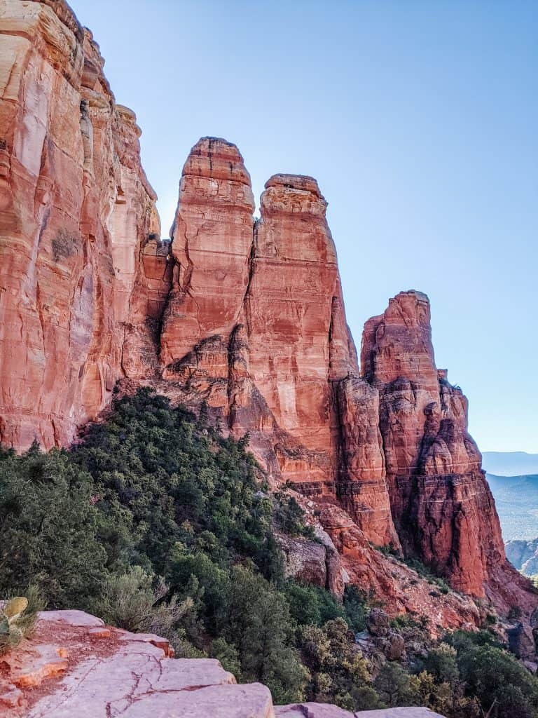 The towering red rock formations in Sedona, known for their connection to the Sedona vortex, with lush greenery at their base and a clear blue sky overhead.