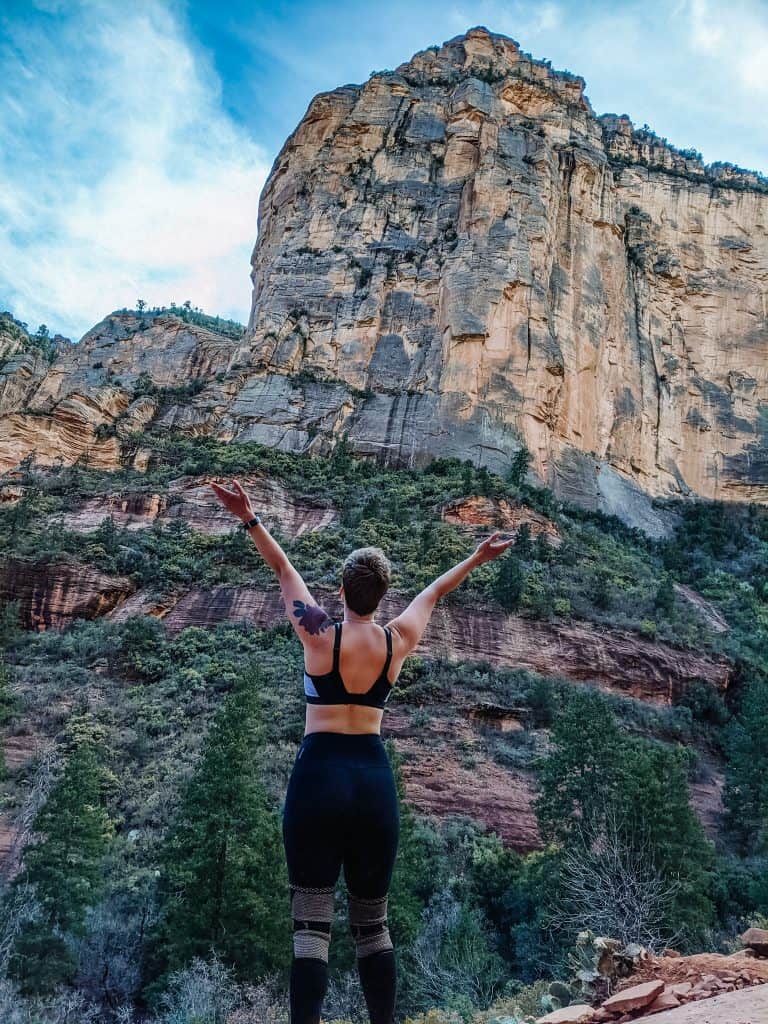 I am standing with my arms raised, facing a towering red rock cliff at the Sedona vortex, surrounded by lush green trees and under a vibrant blue sky.