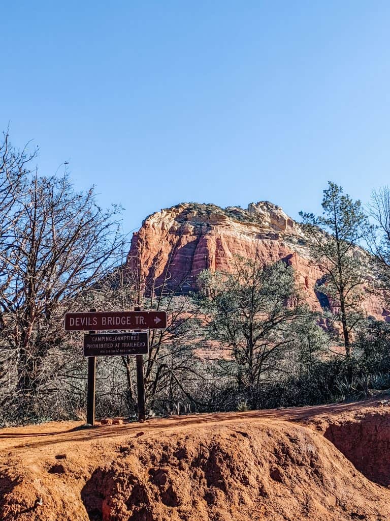 A sign marking the entrance to the Devil's Bridge Trail with the majestic red rock formations of the Sedona vortex in the background, under a clear blue sky.