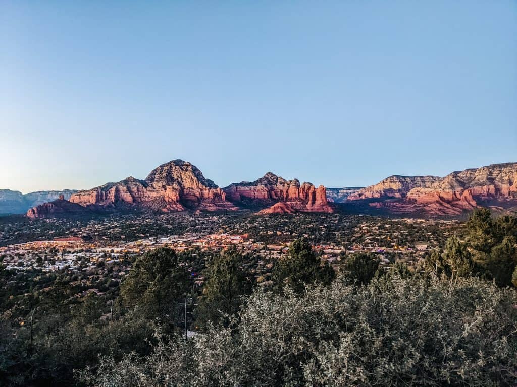 The stunning red rock formations of Sedona at sunset, with the city nestled below the towering cliffs. The landscape is bathed in warm, golden light, enhancing the natural beauty of the area, known for its Sedona vortex sites, which many believe have spiritual energy.