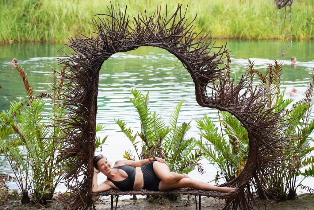 I am lying on a heart-shaped wicker bench beside the clear, green water of Cenote Tulum, surrounded by lush tropical plants, enjoying the serene and natural beauty of the area.