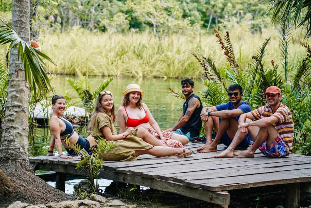 You can see me and my friends sitting on a wooden dock by the water at Cenote Tulum. We are all smiling and enjoying the peaceful, natural surroundings, with lush greenery and clear water providing a perfect backdrop for our relaxing day.