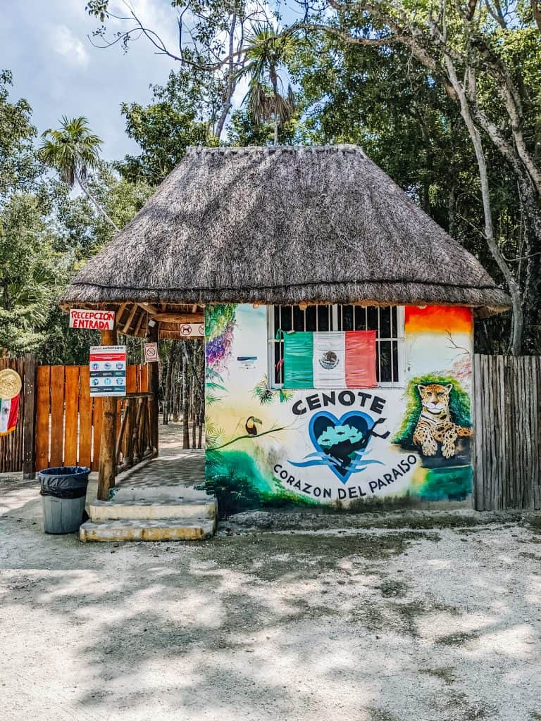 The colorful entrance to Cenote Tulum features a straw roof hut adorned with murals, including a heart-shaped cenote logo and a jaguar.