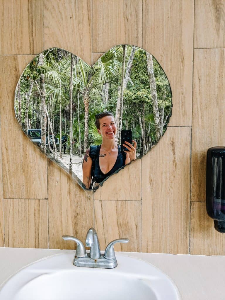 I'm taking a selfie in a heart-shaped mirror, mounted above a sink, with lush greenery visible in the reflection. This creative mirror is found in the restroom area at Cenote Tulum, adding a charming touch to the amenities at this beautiful natural attraction.