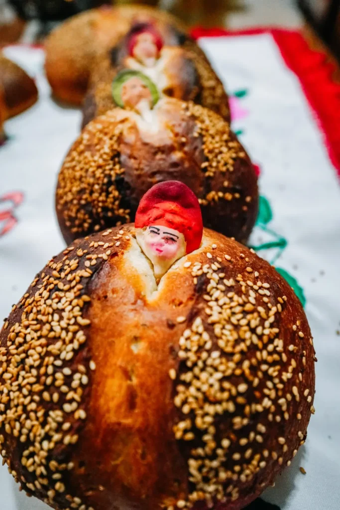 A traditional Mexican bread called "pan de muerto," often prepared for the Day of the Dead Mexico celebrations. The bread is adorned with colorful, small figurines peeking out from the top, surrounded by sesame seeds.