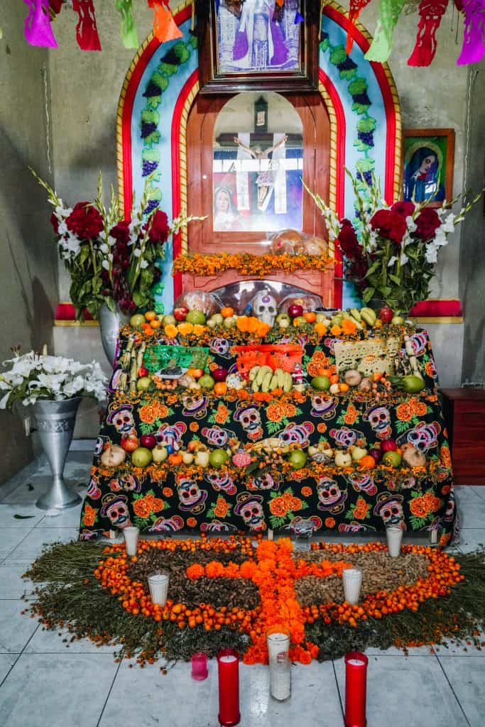 A beautifully decorated Day of the Dead altar in Mexico, adorned with marigold flowers, sugar skulls, candles, and various offerings. The altar is covered with a colorful cloth and framed with bright paper decorations and flowers, creating a vibrant and sacred space to honor the deceased.