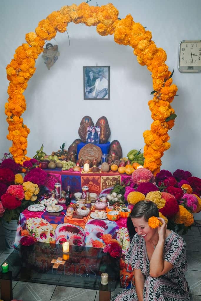 Kneeling beside a beautifully decorated Day of the Dead Mexico altar, which is adorned with vibrant marigold arches, colorful flowers, and various offerings to honor the departed.