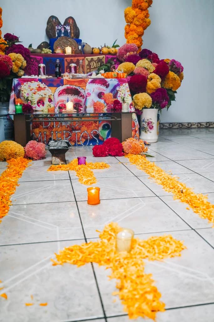 A Day of the Dead Mexico altar with a path of marigold petals leading to it. The marigold petals form a cross on the tiled floor, guiding the spirits to the altar. The altar itself is decorated with vibrant flowers, candles, and offerings, creating a colorful and sacred space for honoring the deceased.