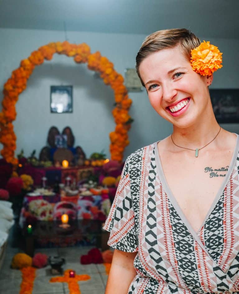 Me smiling with a bright marigold flower in my hair, standing in front of a beautifully decorated Day of the Dead altar in Mexico. The altar, adorned with vibrant marigold arches, candles, and offerings, reflects the rich traditions of honoring deceased loved ones.