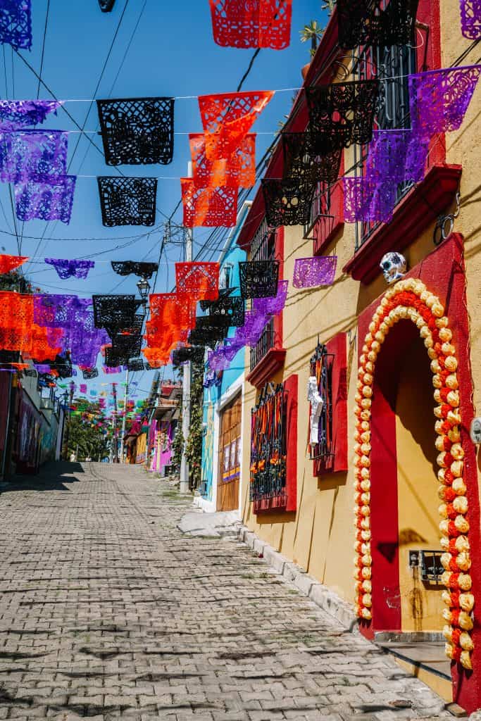 A vibrant street in Mexico is adorned with colorful paper flags in purple, red, and orange for the Day of the Dead celebrations. The cobblestone road is lined with buildings, one of which has a marigold arch framing the door, symbolizing the path for the spirits to follow.