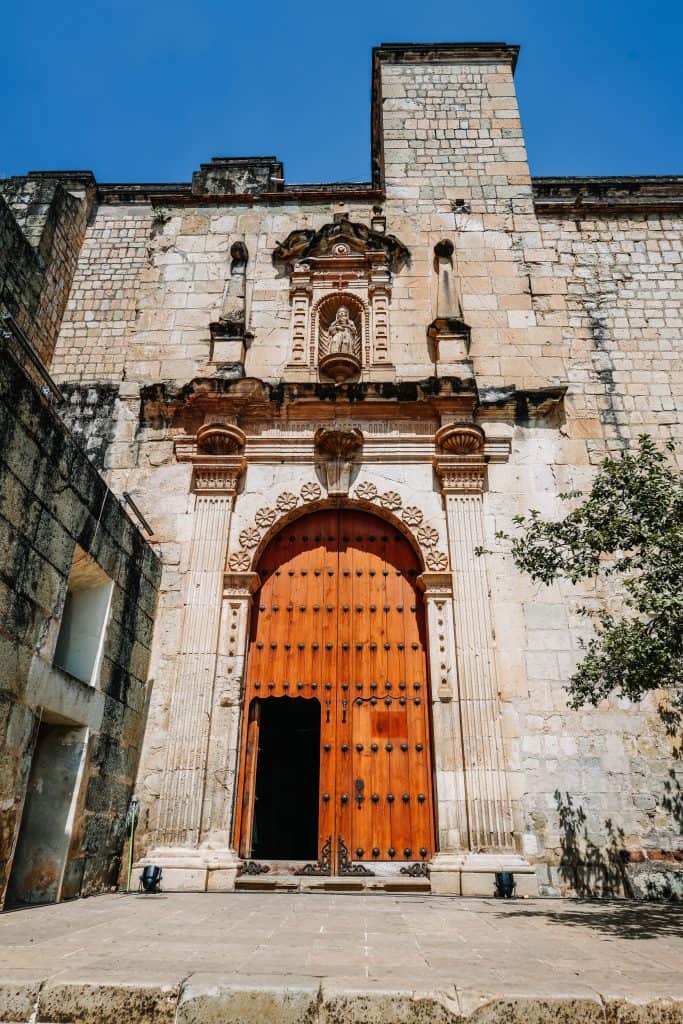 A grand, ancient church entrance with a massive wooden door, adorned with intricate carvings, standing tall against the blue sky. The historical architecture is a significant part of the Day of the Dead Mexico celebrations, where locals gather to honor their ancestors.