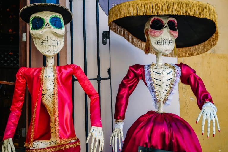 Two colorful skeleton figures dressed in traditional attire, often seen during Day of the Dead celebrations in Mexico. One skeleton wears a vibrant red dress with a wide-brimmed hat, while the other sports a red suit with complex designs.