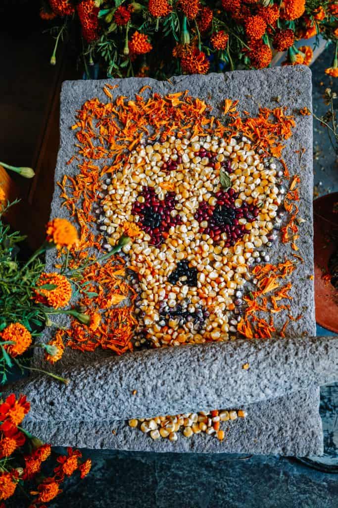 A creative sand painting for Day of the Dead in Mexico, featuring a skull made from yellow corn kernels and marigold petals, with red flowers as the eyes.