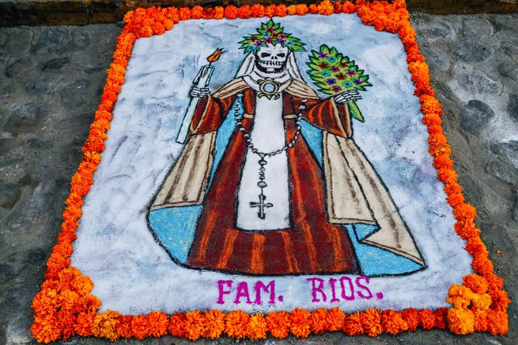 A detailed sand painting for the Day of the Dead in Mexico features a skeleton dressed in traditional clothing, holding a bouquet and candle, surrounded by bright marigold flowers with the name "Fam. Rios" written at the bottom.