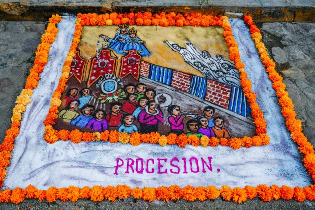 A vibrant sand painting created for the Day of the Dead in Mexico, illustrating a procession. The artwork is bordered with bright marigold flowers and features a crowd of people participating in a parade. The figures are dressed in colorful clothing, and at the center, there is a detailed depiction of a religious icon being carried on a decorated platform. The word "PROCESION" is written at the bottom, emphasizing the theme of the painting.