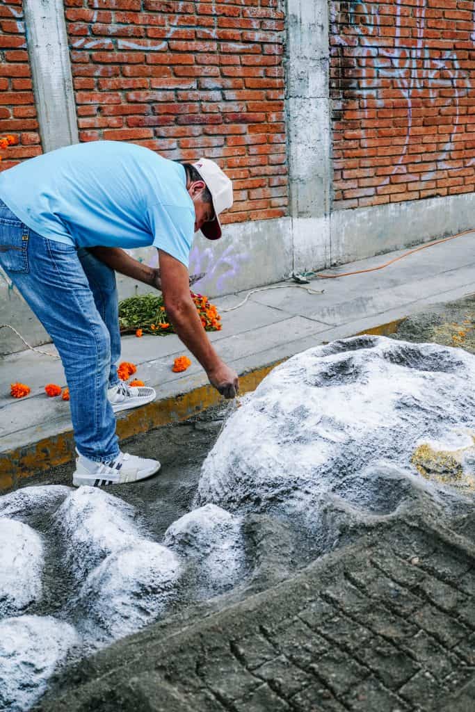 A man wearing a blue shirt and white cap creates a sand painting on the sidewalk for Day of the Dead in Mexico, a tradition that involves intricate artwork to honor the deceased.