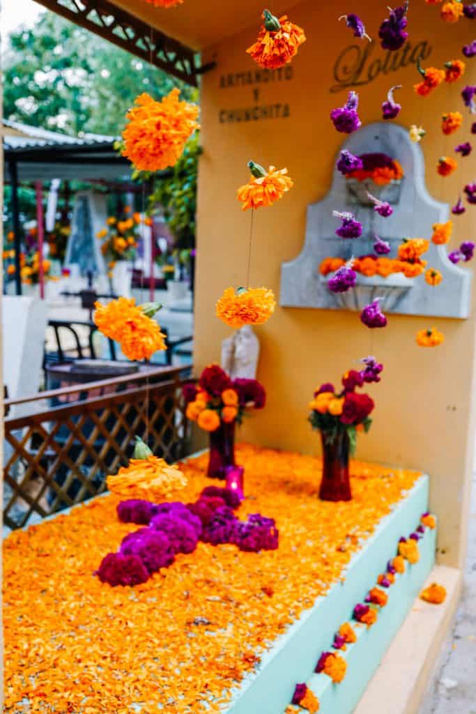 A grave decorated for the Day of the Dead in Mexico is covered with a blanket of bright marigold petals and adorned with vibrant purple and yellow flowers in vases, creating a colorful and heartfelt tribute.