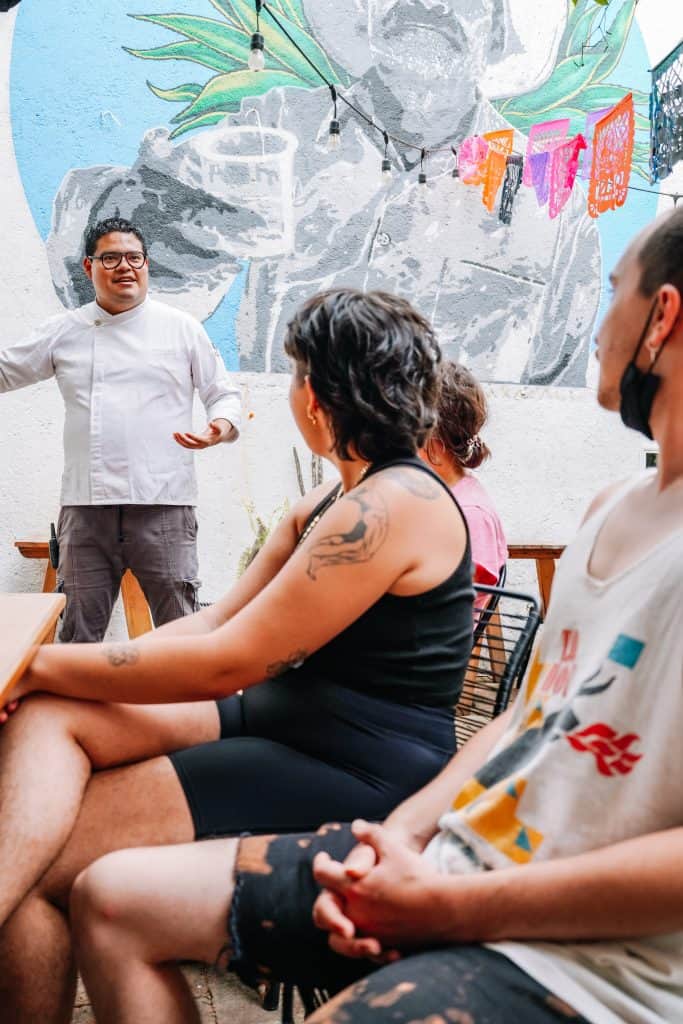 A chef is engaging with a small group of people, likely sharing insights about traditional Oaxacan cuisine at an Oaxaca restaurant. The mural in the background adds a colorful and cultural touch to the setting, while the attentive expressions of the listeners reflect their interest in learning about the rich culinary heritage of Oaxaca.