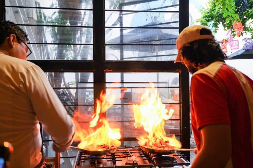 A chef and an assistant work together at an Oaxaca restaurant, both intensely focused on pans with flames leaping high, suggesting a vibrant cooking session with rich, flavorful dishes in the making. The bright fire contrasts with the soft natural light coming through the windows.