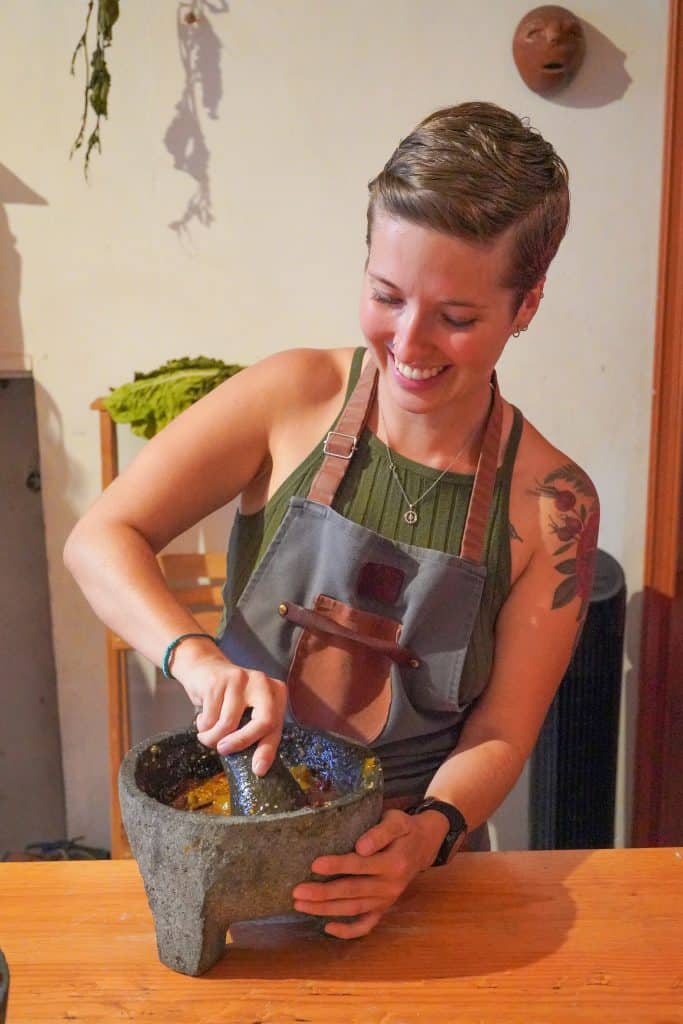 Joyfully grinding ingredients with a traditional stone mortar and pestle, preparing food for Day of the Dead celebrations in Mexico, while wearing an apron and standing at a wooden counter, fully immersed in the cultural experience.