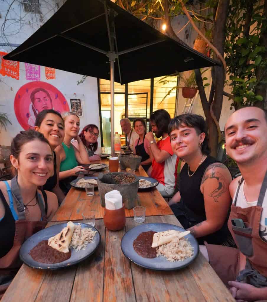 My friends is seated around a wooden table at an Oaxaca restaurant, enjoying a meal that includes plates of mole with rice and tortillas, under the soft glow of string lights and a large umbrella.