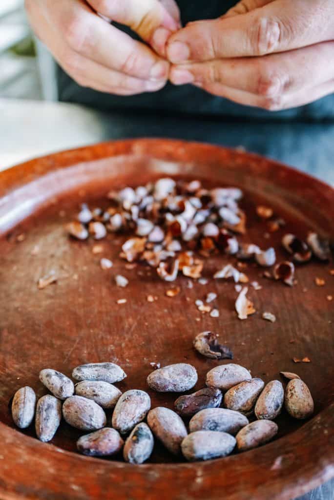 Hands are seen carefully peeling the shells off cacao beans on a wooden plate, a practice that reflects the meticulous preparation process often found in a traditional Oaxaca restaurant.