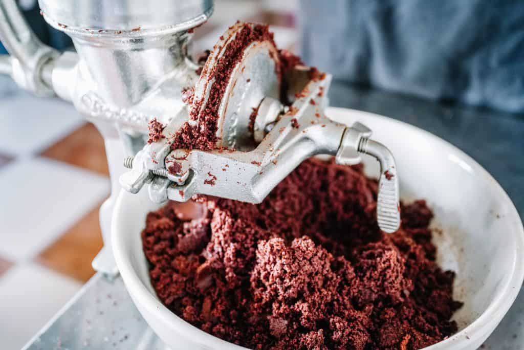 A hand-crank grinder is being used to grind cacao beans into a coarse paste, a traditional method often seen in a Oaxaca restaurant. The close-up view captures the gritty texture of the cacao and details of the metal grinder.