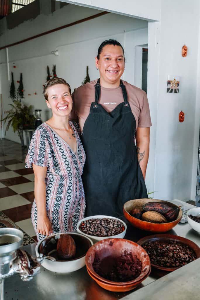 My friend and I are standing in a cozy Oaxaca restaurant kitchen, smiling next to a table filled with bowls of cacao beans, cacao pods, and ground chocolate, ready to be made into traditional Mexican chocolate.