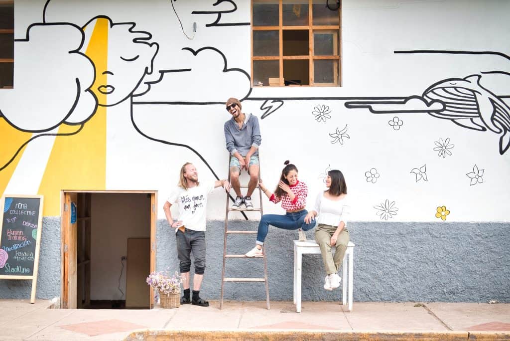 A group of friends are casually hanging out in front of a mural outside one of the vegan restaurants in Sacred Valley, Peru. The mural features playful and artistic black-and-white drawings with a bold yellow stripe. The friends are sitting and standing on various outdoor furniture, smiling and enjoying their time together