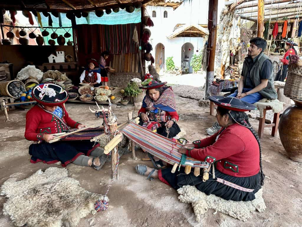 Women dressed in traditional Peruvian attire are seated on the ground, weaving colorful textiles using ancient techniques. They are under a rustic shelter, surrounded by vibrant crafts and materials, while a few onlookers observe the process.