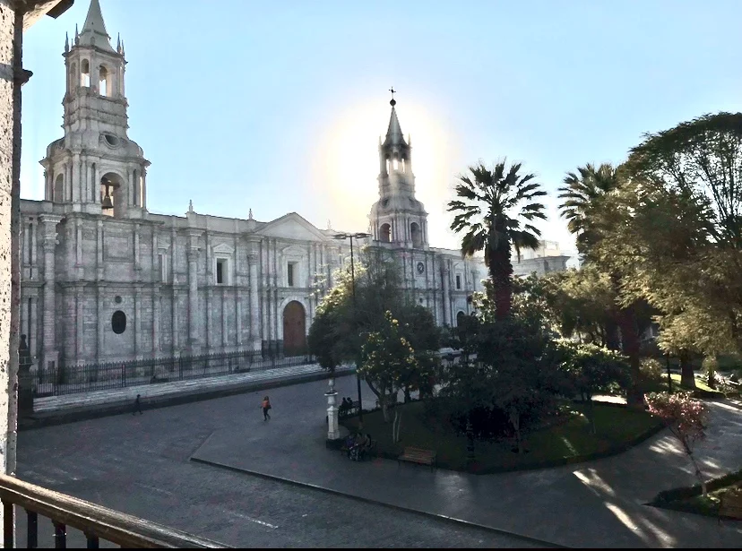 One of the notable places to visit in Peru, the majestic Arequipa Cathedral stands tall with its twin bell towers gleaming in the morning sun. The plaza in front of the cathedral is lined with lush trees and benches. A lone figure can be seen walking, adding a sense of quiet. The cathedral's white stone front contrasts beautifully with the clear blue sky, making it a striking landmark in Arequipa.