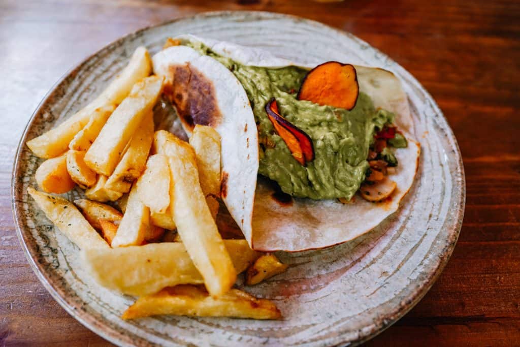 A delicious vegan meal from a restaurant in Sacred Valley, Peru is displayed. The plate features a well-stuffed taco with a generous portion of guacamole, paired with crispy potato wedges, all presented on a rustic ceramic