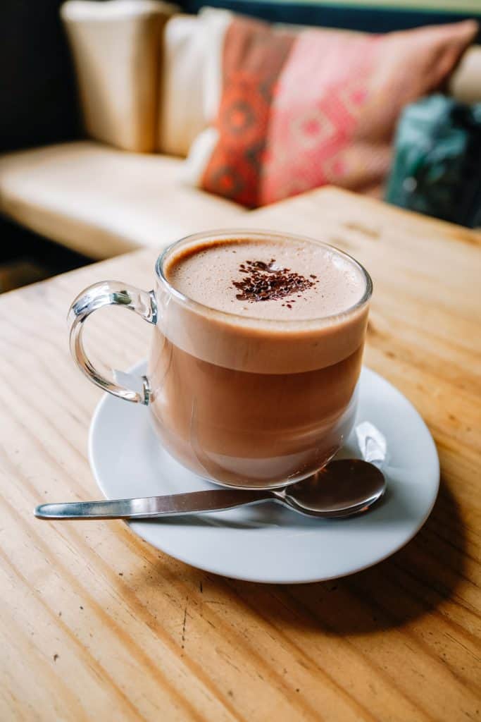 A cozy corner in a vegan restaurant in Sacred Valley, Peru, with a warm, frothy cup of vegan hot chocolate served in a clear glass mug, resting on a white saucer with a silver spoon beside it. The background features soft, colorful pillows, enhancing the inviting and relaxed atmosphere.