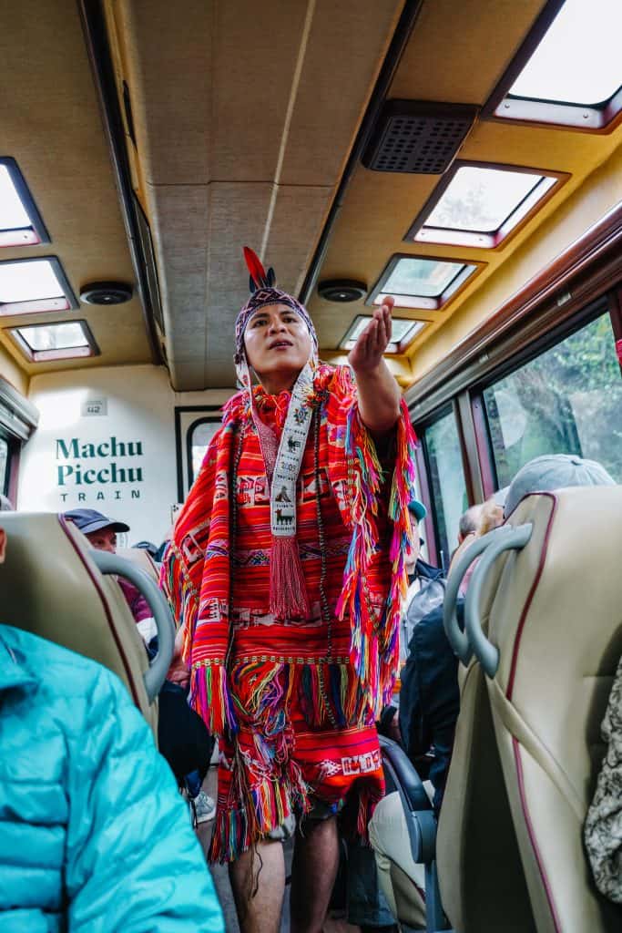 A man dressed in traditional colorful Peruvian attire performs a cultural dance inside the Machu Picchu train, enhancing the journey to the ancient site. Experiencing such cultural performances is one of the many engaging things to do at Machu Picchu.