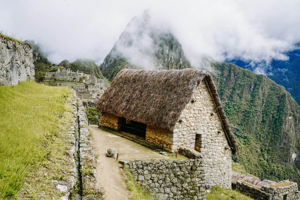 A thatched stone house sits on a narrow path overlooking the misty green mountains and ancient ruins of Machu Picchu. One of the essential things to do at Machu Picchu is exploring these well-preserved structures.