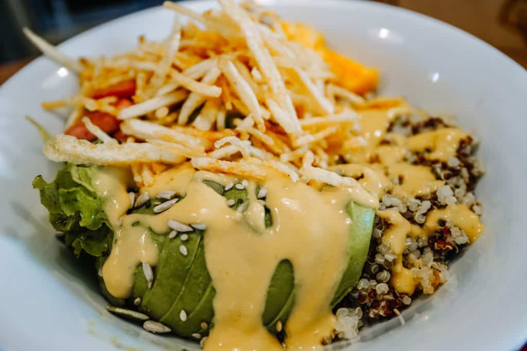 A hearty vegan bowl from a restaurant in Sacred Valley, Peru, filled with quinoa, fresh avocado, shredded carrots, and leafy greens, all drizzled with a creamy, tangy dressing and topped with crispy potato sticks.