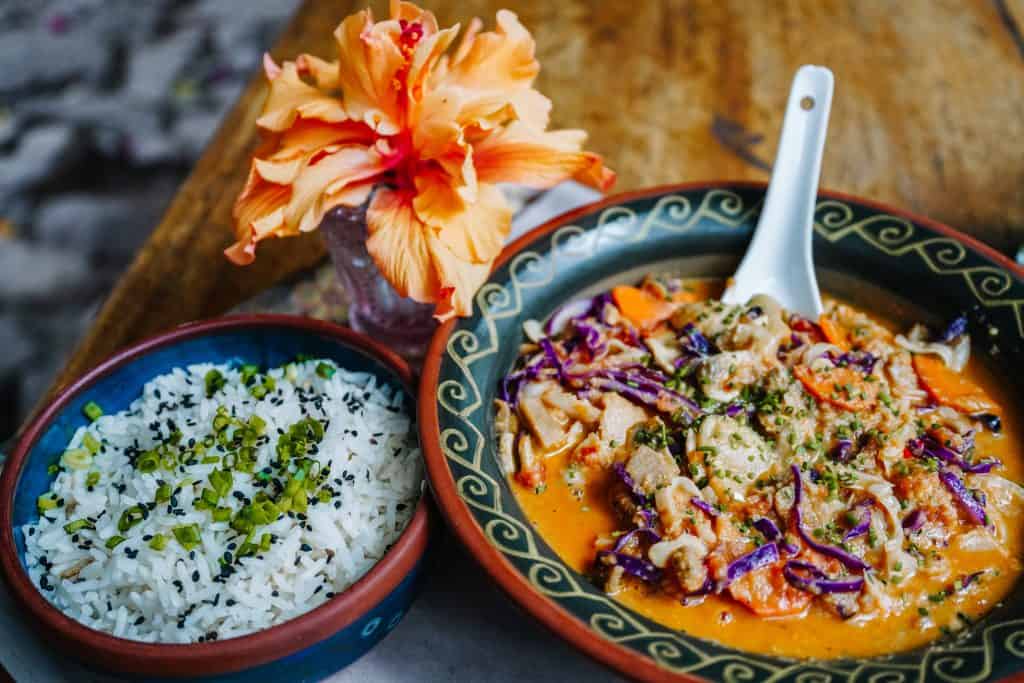 A delicious vegan dish served at a vegan restaurant in Sacred Valley, Peru. The meal consists of a hearty bowl of colorful vegetables in a rich, creamy curry sauce, garnished with fresh herbs. Accompanying the main dish is a side of perfectly cooked rice sprinkled with sesame seeds and chopped greens. A bright orange flower in a small vase adds a touch of natural beauty to the rustic wooden table, enhancing the overall dining experience.