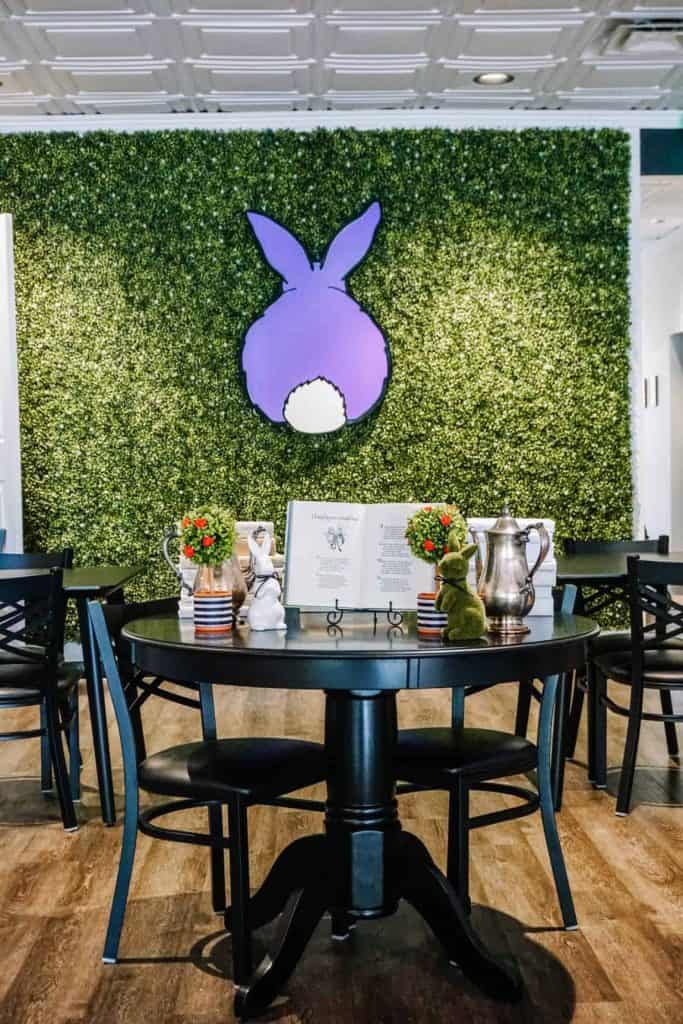 I captured this charming setup featuring a black round table with elegant chairs. The table is adorned with small vases of flowers and tea accessories, set against a backdrop of a lush green wall with a whimsical purple bunny tail art piece at Drink Me Tea Room in Arizona.