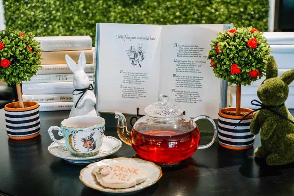 At Drink Me Tea Room, I captured this playful setup featuring a teapot filled with vibrant red tea, a teacup, and a "Drink Me" cookie. The table is adorned with charming decorations, including a white rabbit figurine, a small book of poems, and two topiary trees, creating a magical and inviting atmosphere.