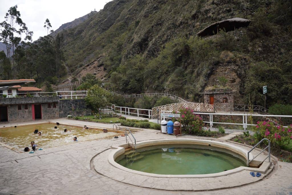 A hot springs area nestled in the Peruvian mountains, where visitors can be seen enjoying the warm, waters of the pools surrounded by lush greenery and rugged hills. The inviting circular pool in the foreground contrasts with the larger, more rustic pool in the background.