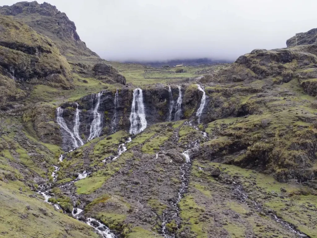 A stunning waterfall cascades down the rocky, green mountainside in the Andes, a must-see among places to visit in Peru, with misty clouds hovering above the rugged landscape.