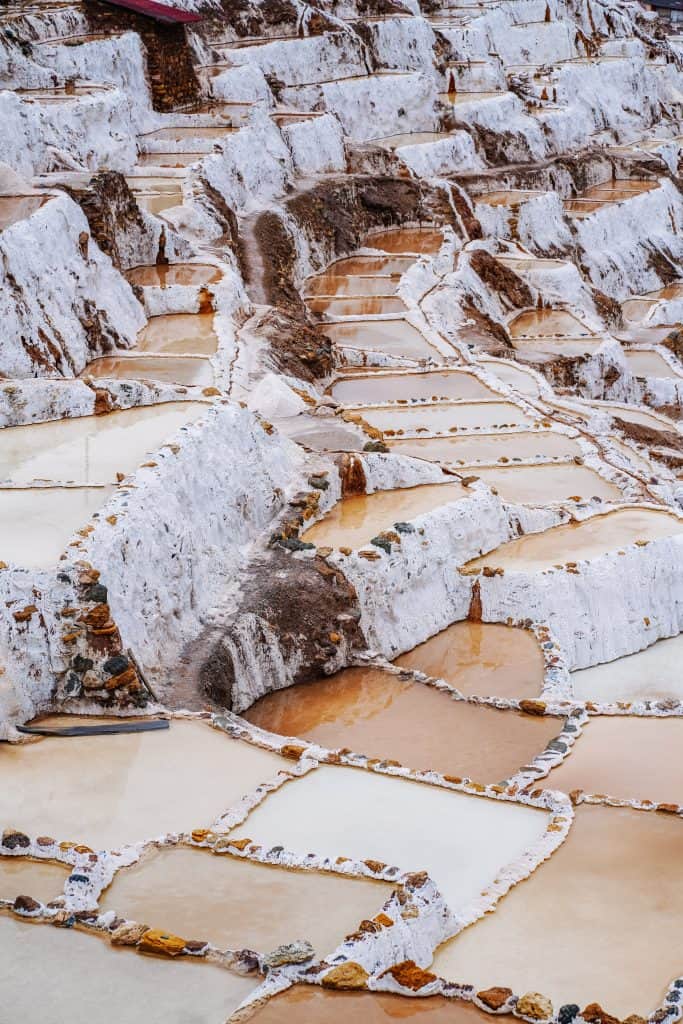 The complex salt ponds of Maras, one of the fascinating places to visit in Peru. The terraced ponds, filled with varying levels of water and salt, create a striking mosaic pattern against the rugged Andean landscape. The white salt crusts contrasting with the brown earth give the scene a surreal.